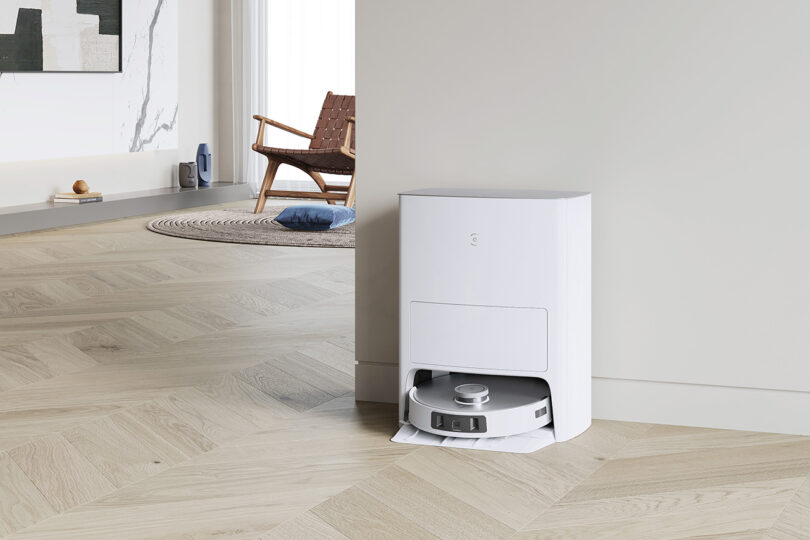 Robot vacuum recharging or emptying in its white base station set against a living room wall, with modern armchair in the background.