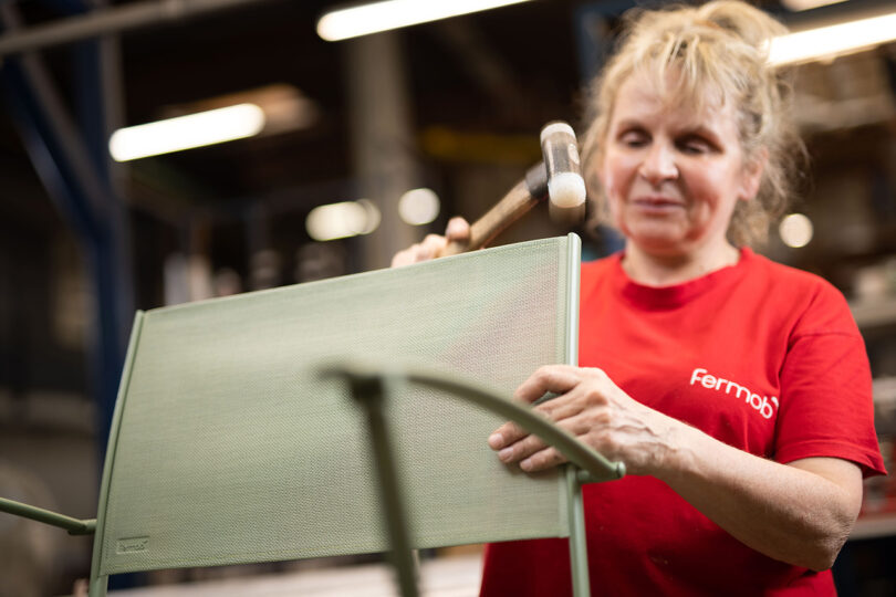 Woman in red t-shirt hammering back support of mesh and metal frame Fermob chair into place within factory setting.