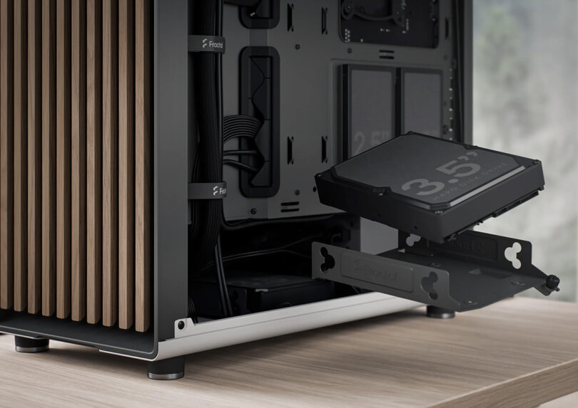 Black PC tower case with on left side of a wood desk seen from rear with left side panel open, revealing two 2.5-inch solid state drives and one 3.5-inch drive being slotted into case using a caddy.