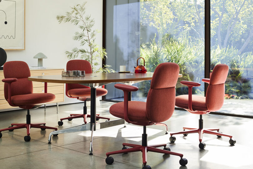 Four red Asari mid-height tasks chairs in office conference room setting around oval table with large window looking out into garden.