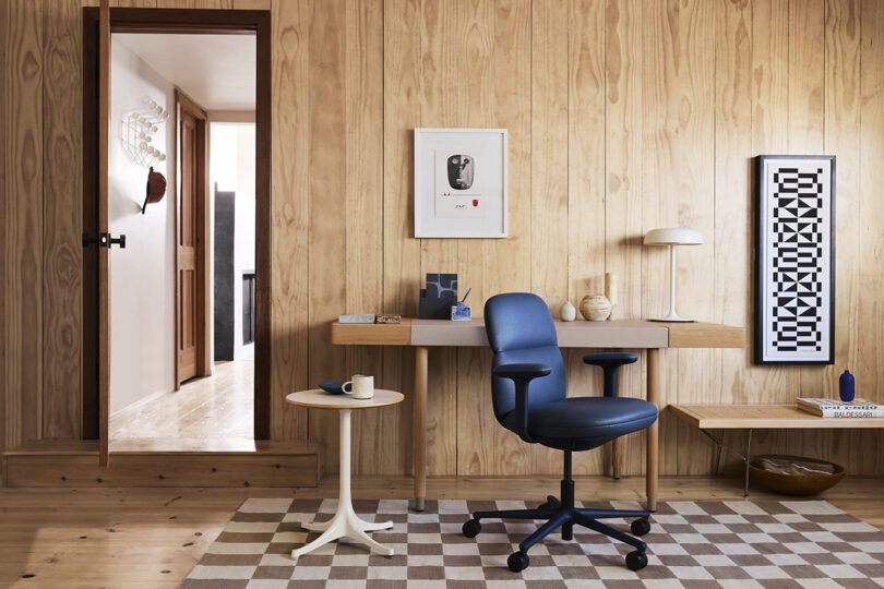 صندلی کار آساری به رنگ مشکی در اتاق نشیمن با محیط دفتر خانه با دیوارهای پانل چوبی و فرش شطرنجی با درب باز به سمت چپ.