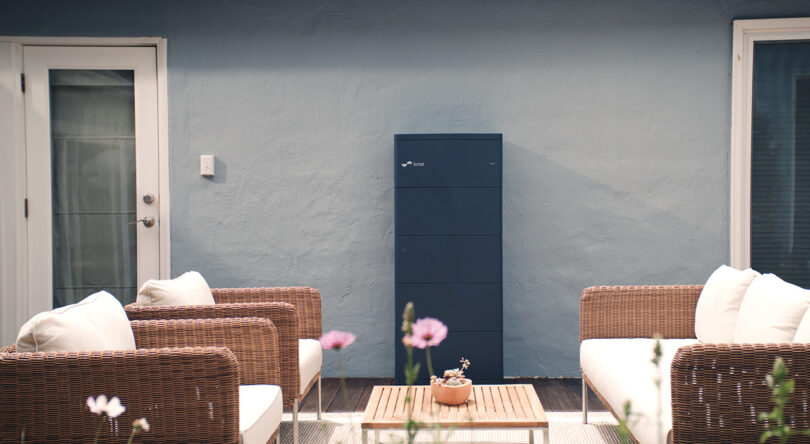 Lunar Energy battery system in dark blue set against a white exterior wall of a home, with outdoor wicker seating and table in the foreground