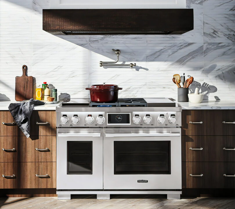 Signature Kitchen Suite 48 اینچی دوگانه سوز با هود با کابینت چوبی و پشتی کاشی طرح مرمر نشان داده شده است.