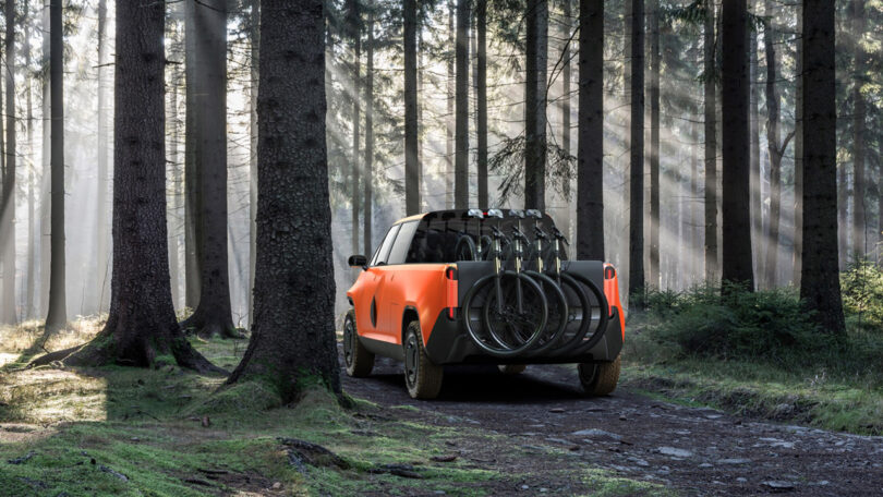 Orange TELO electric truck parked in forest from rear view angle, the back truck bed loaded with four mountain bikes with front wheels overhanging from bed door.