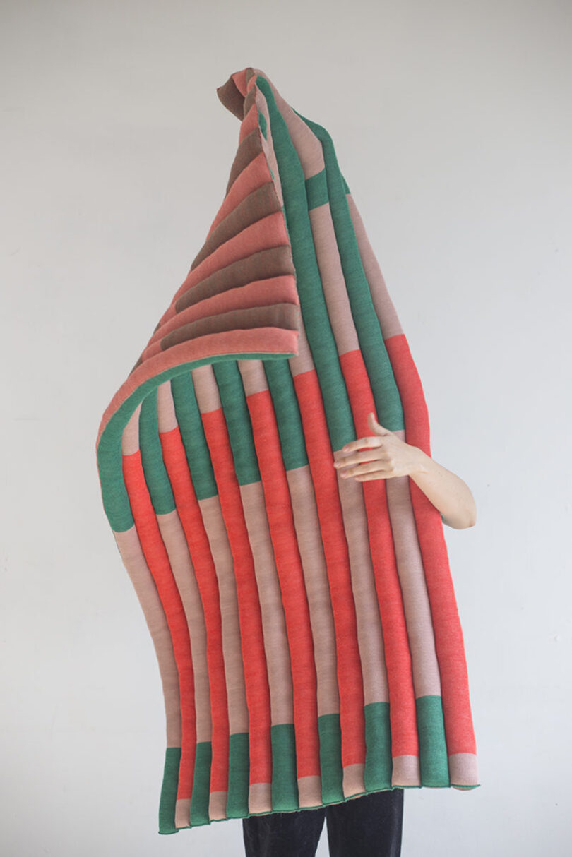 light-skinned person holding up a large piece of red, green, and tan woven art