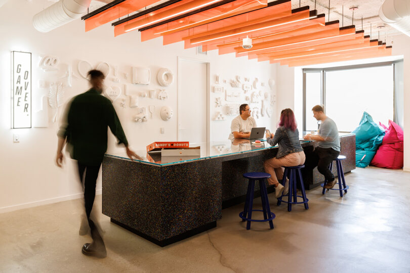 Wide view of long glass table following a long white wall displaying a mix of emojis and other icon-based graphics protruding from the surface, alongside a "Game Over" sign glowing to the left. Orange bannered lighting is overhead.