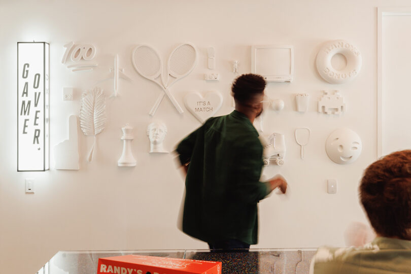 Back view of man in green long sleeve shirt looking at white wall displaying a mix of emojis and other icon-based graphics protruding from the surface, alongside a "Game Over" sign glowing to the left.