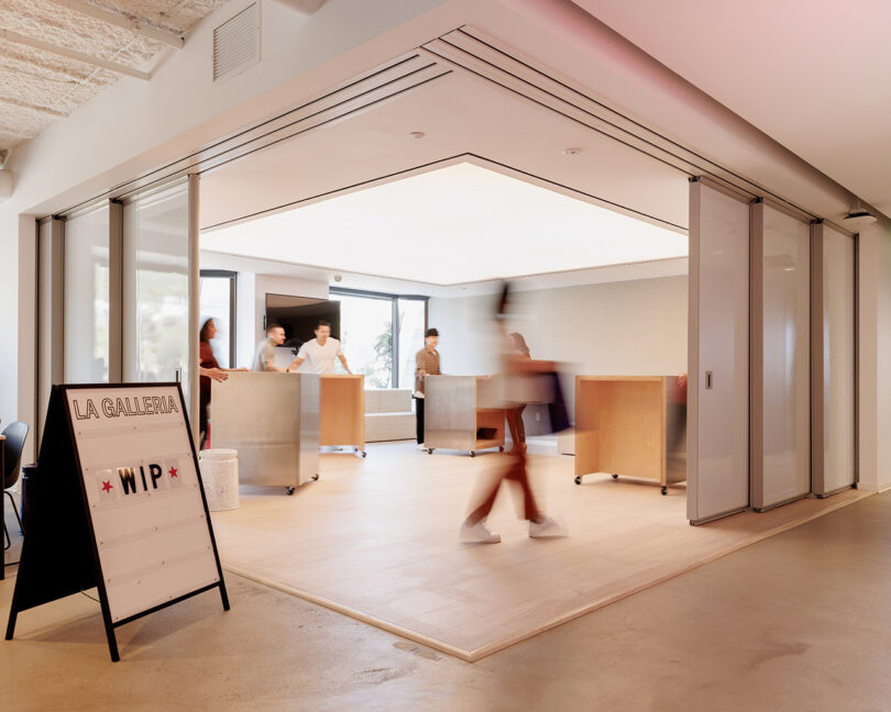 Room with two sides of corner sliding doors open with a "La Galleria" sign outside with "WIP" displayed on it; people inside are moving standing desks on wheels.