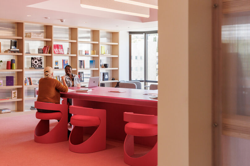 Two women seated at a hot pink desk and chairs near floor to ceiling bookshelves inside Tinder headquarters.