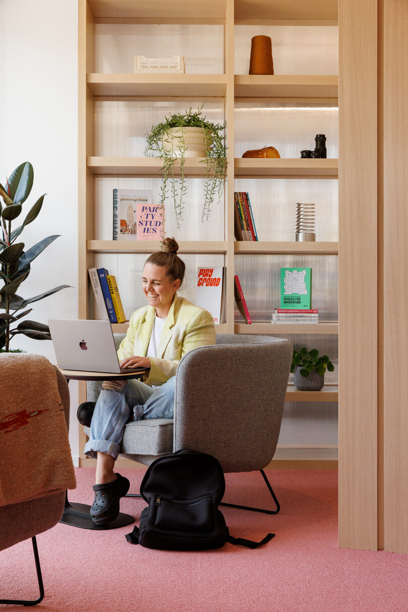 Young woman seated and laughing, looking at her laptop seated in a gray armchair with bookshelf in background and backpack on the floor.