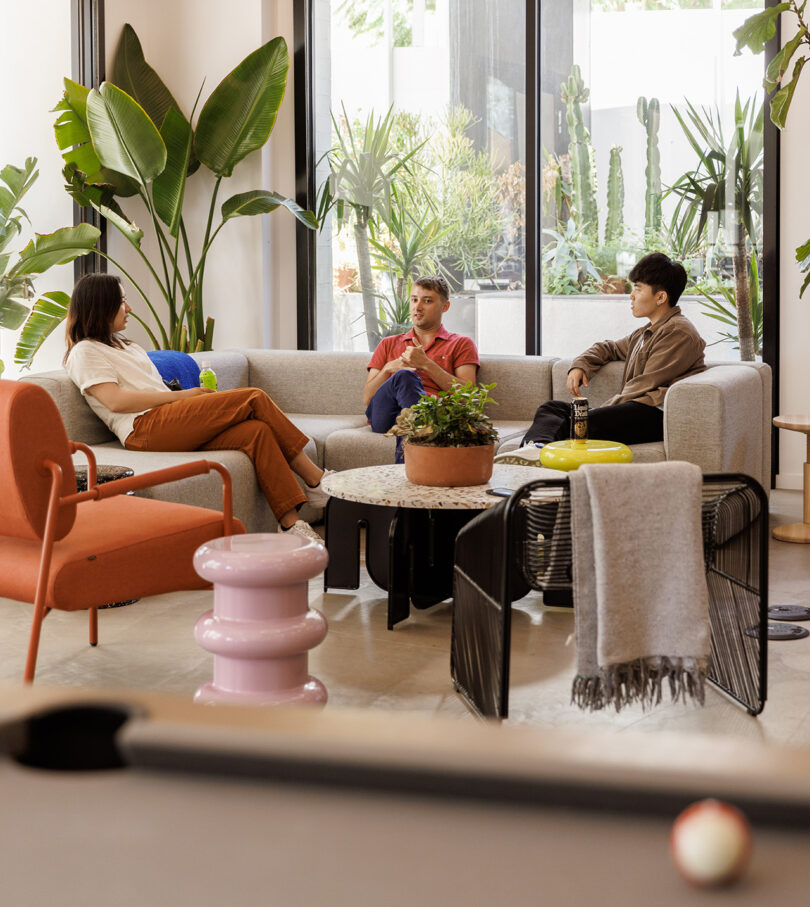 One young woman seated with two young men across sectional sofa conversing in Tinder HQ surrounded by palm plants.