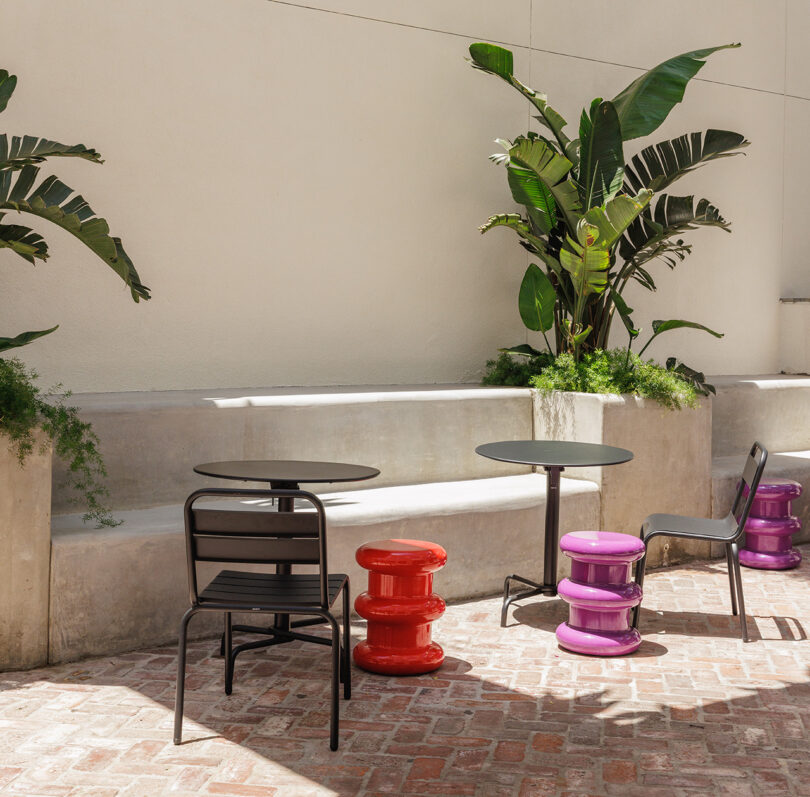 Outdoor seating area with brick floor, black metal chairs and tables, alongside a trio of red and light purple stools surrounded by large palm leaf plants.