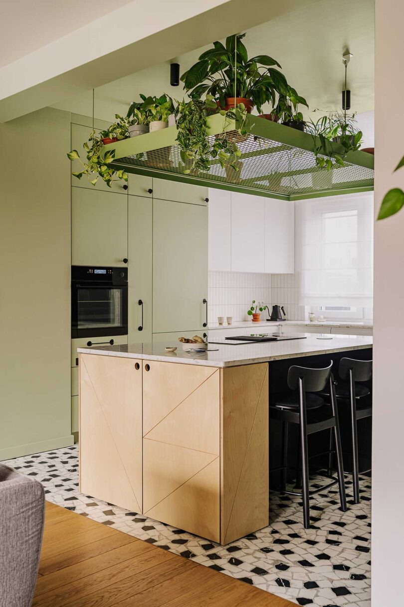 نمای زاویه دار آشپزخانه مدرن با ترکیبی از کابینت های مینیمالیستی به رنگ سفید و سبز مریم گلی و جزیره چوبی روشن با گیاهان آویزان در بالا