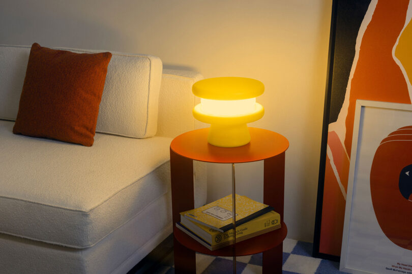Yellow macaron shaped table lamp set on orange-red circular side table next to off white sofa.