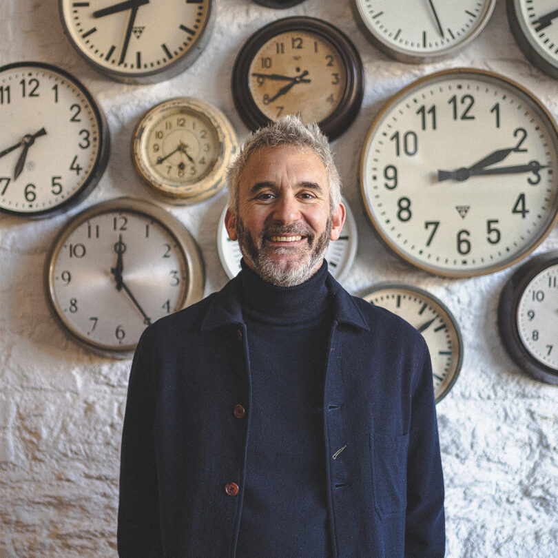 Chris Miller, a white man in his 50s, stands in front of a wall of clocks