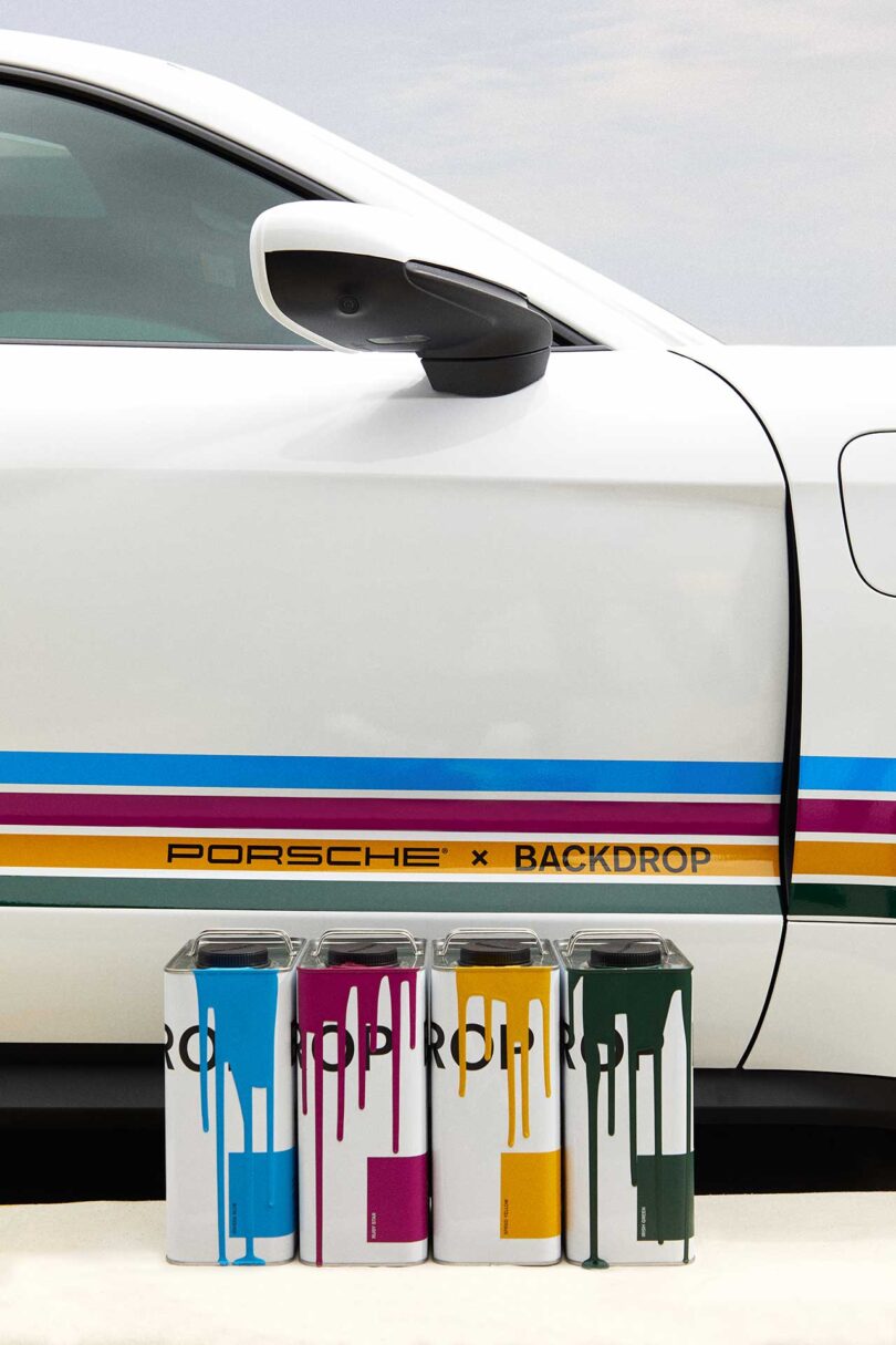 partial closeup view of white Porsche with four colorful stripes of paint running down the side with four cans of paint in front