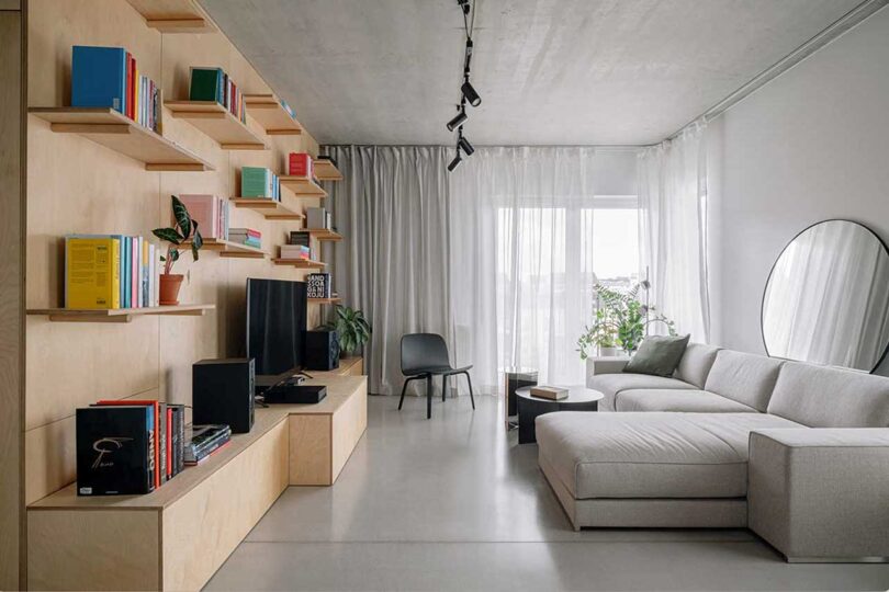 view of modern living room with light wood box structure within an apartment holding shelves of books and TV opposite a light grey sectional sofa