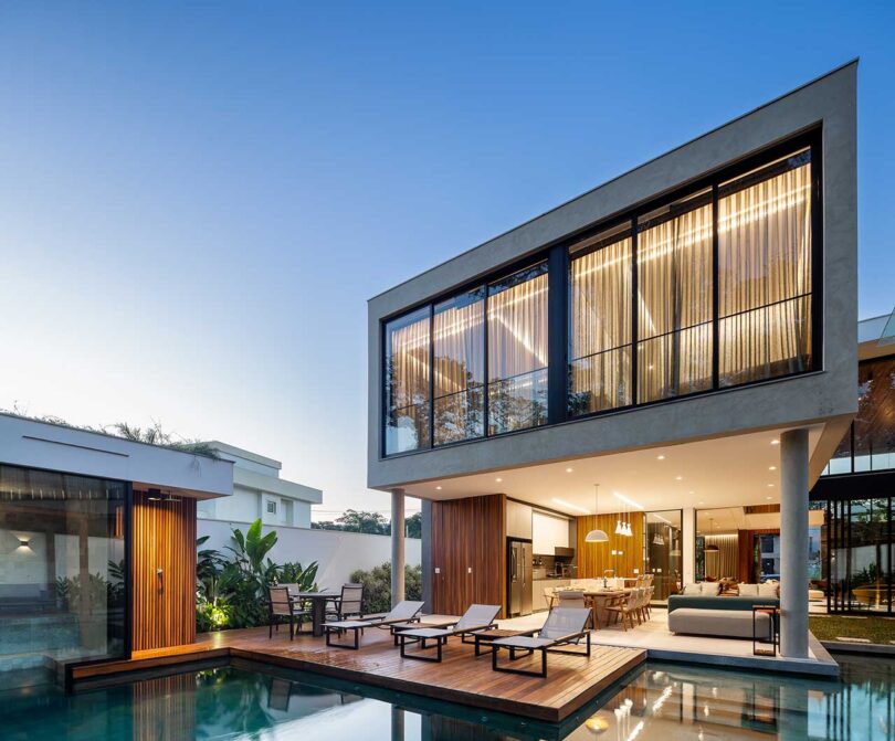 angled exterior view of modern home's rear at sundown with lights on and views of the pool