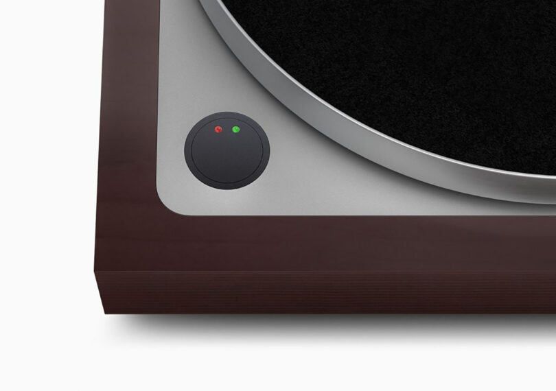 Detail of the corner of the turntable showing the circular, machined-from-solid aluminum power switch with red and green dot lights, designed by Jony Ive.
