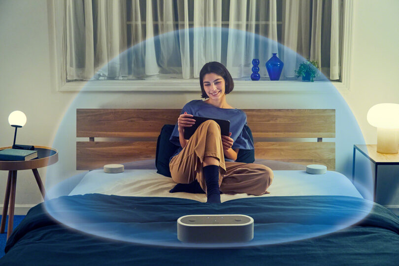 Young woman seated in a bed with blue covers with a Sony HT-AX7 home theater audio speaker system paired with her tablet at the foot of the bed. A illustrated surround sound sphere wraps around her to emulate the immersive sound.