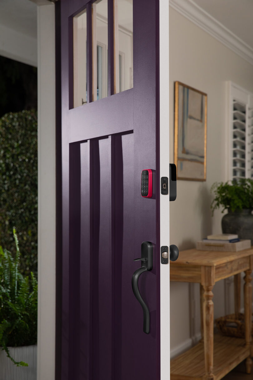 Yale Assure Lock 2 with a Viva Magenta color case installed onto a dark purple front door with black door handle set halfway ajar, illustrating how the smart lock looks from both inside and outside once installed.