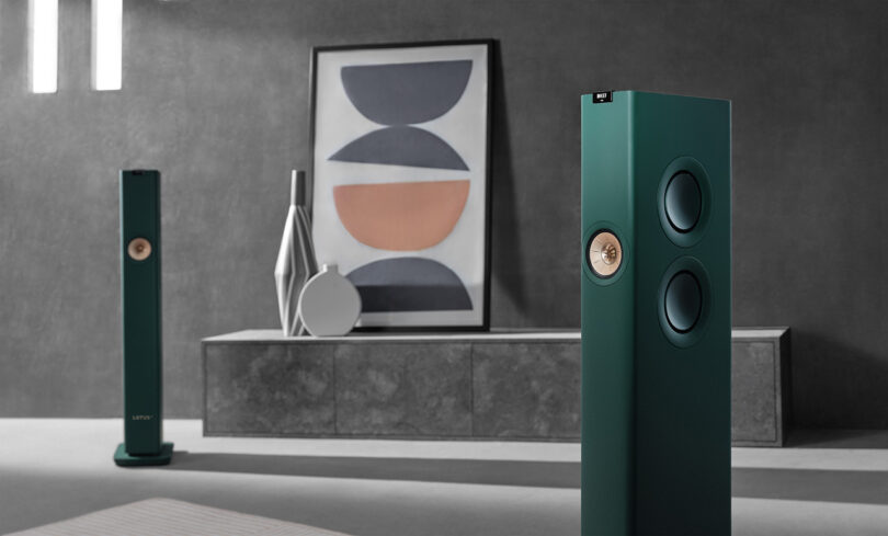 Angled view of KEF LS60 Wireless Lotus Edition Speakers in British Racing Green within modernist concrete wall interior and abstract framed art and vases in background.