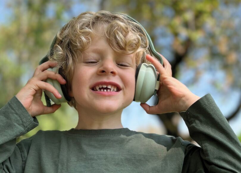 Young boy with curly blond hair wearing and holding each ear cup of the Kibu headphone in light green.