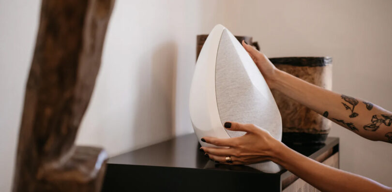 Pantheone Obsidian wireless speaker in white set within an organic modern decor setting on black cabinet with woman's arms reaching to lift it.