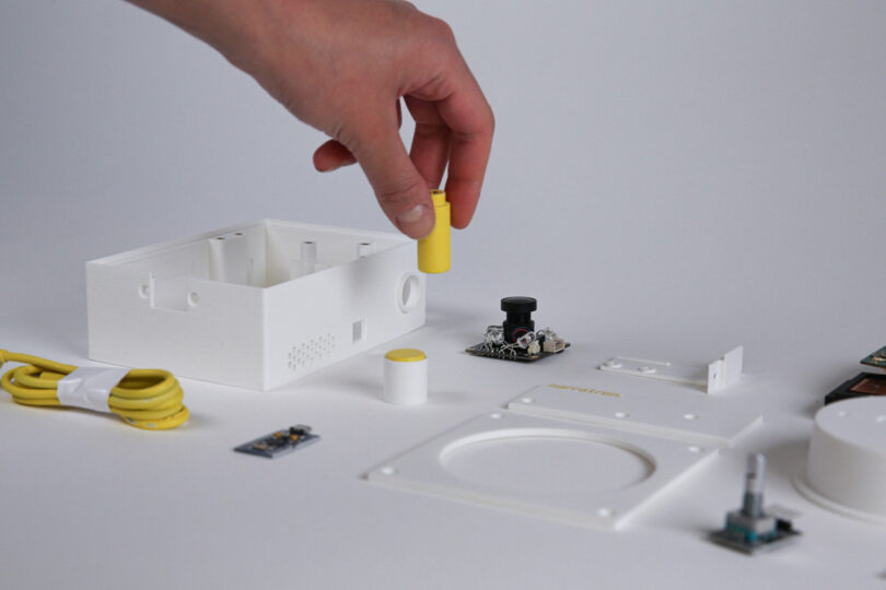 Hand hovering over the disassembled parts of the Narratron projector holding a plastic yellow cylindrical piece.