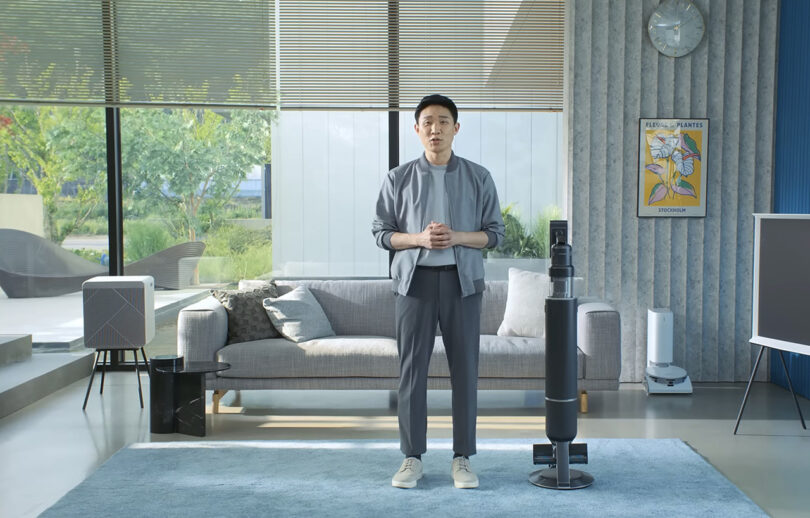 Korean young man in a living room set with modern furnishings and Samsung home appliances, with Samsung Bespoke Jet AI vacuum to his left.