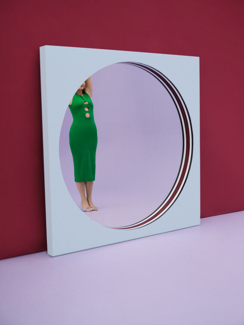 round mirror within large baby blue square frame reflecting a light-skinned woman in a green dress