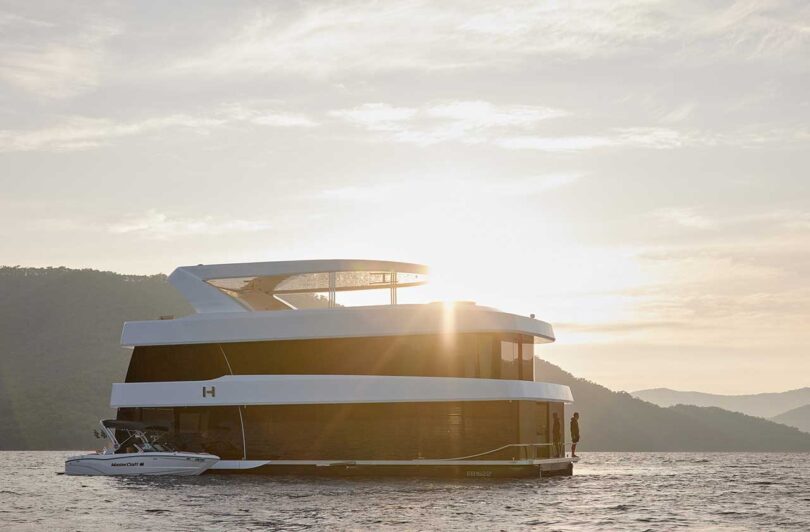 exterior of modern multi level white houseboat with black glass floating on lake with sun setting behind it