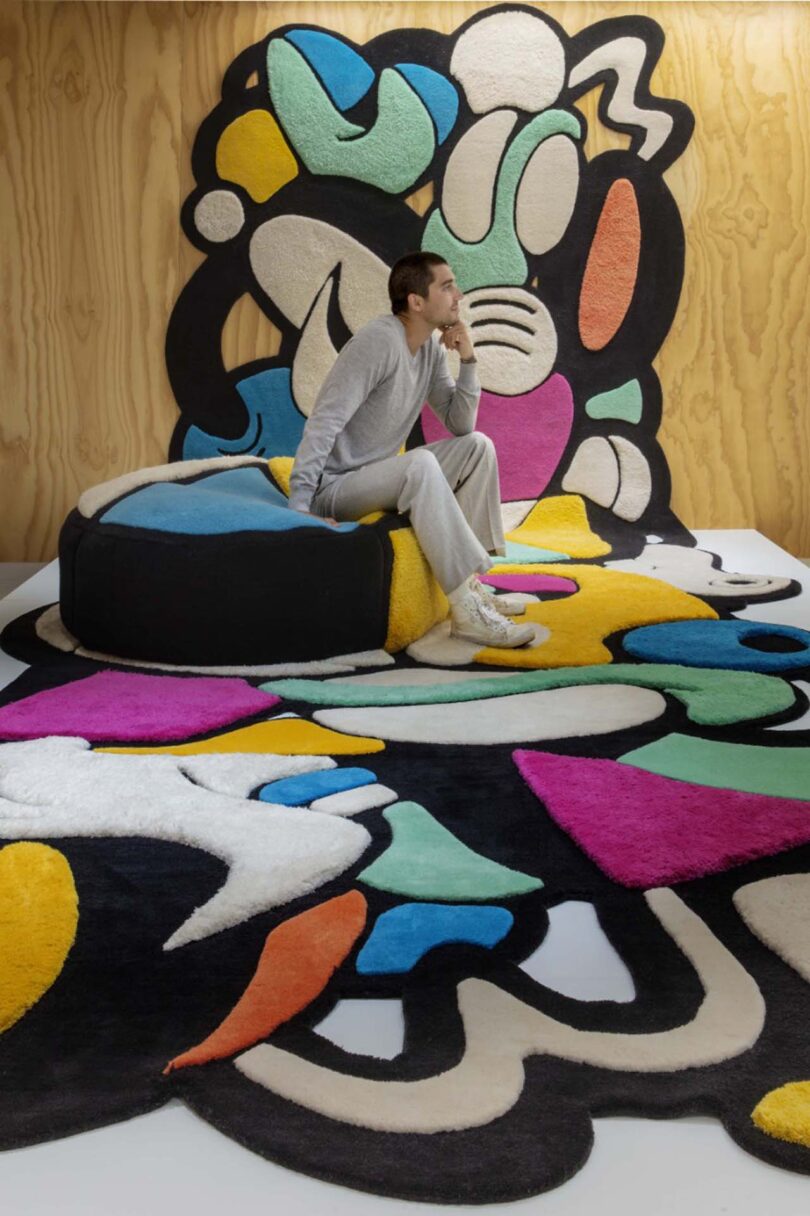 man sitting on a colorful round pouf that's on top of a large colorful rug that flows from floor to ceiling