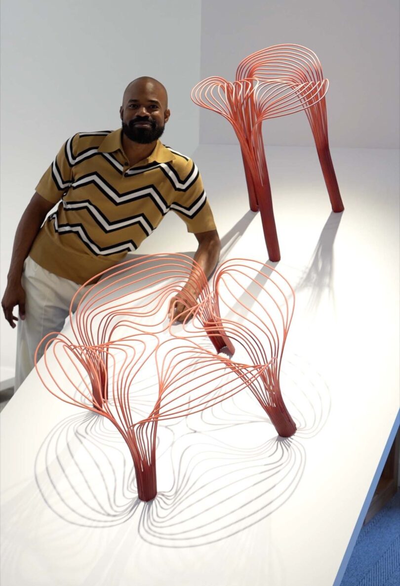 brown-skinned man poses with two pieces of furniture made from red and blue wires