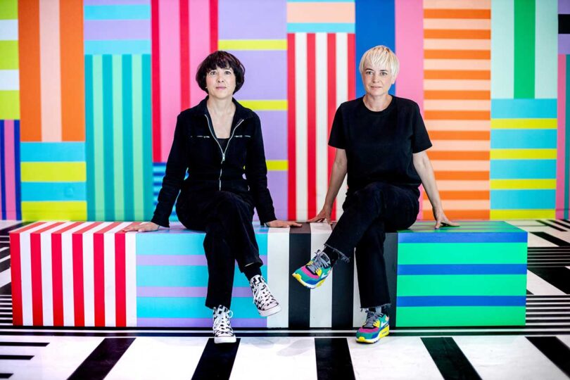 two light-skinned people wearing all black sit in a space space filled with multiple stripe patterns and colors