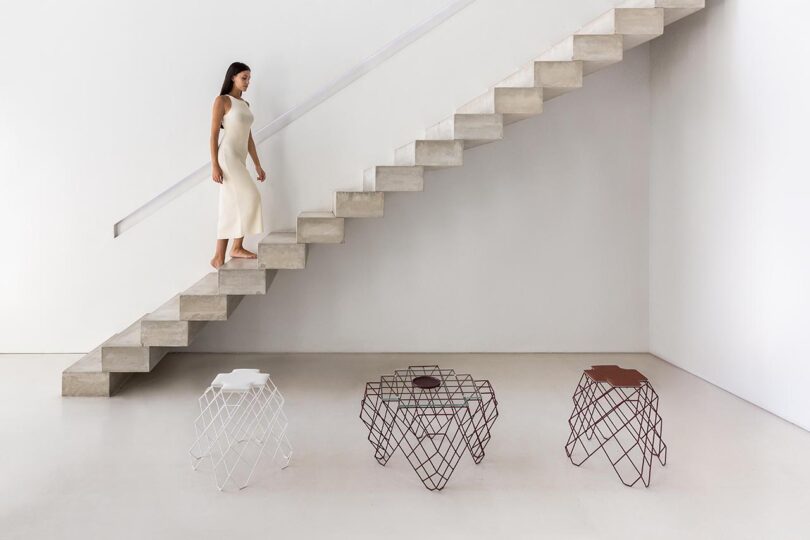 light-skinned woman walking up a tall staircase with three wire work stools/side tables in the foreground