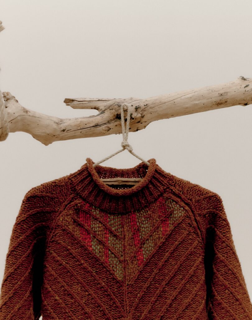 rust colored sweater with threaded pattern sewn at top hanging on branch