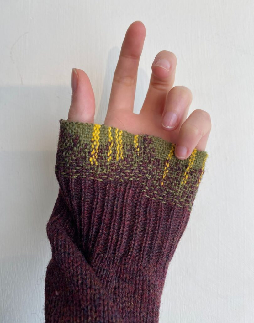 person's hand held up straight with plum colored sweater partially covering hand with green and yellow threaded pattern at sleeve