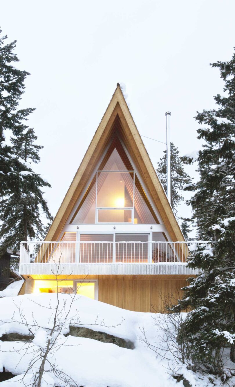 exterior shot of angular a-frame cabin in light wood with white accents surrounding by snowy trees