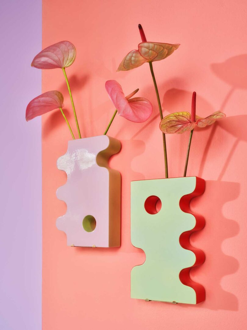 angled closeup view of pink wall with two abstract ceramic vases handing onto wall with flower stems