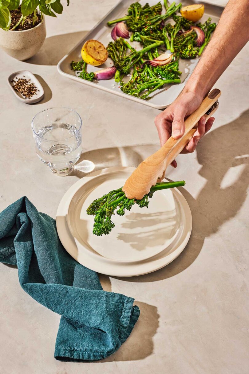 styled shot of dining table with hand extended holding wooden tongs placing greens on plate