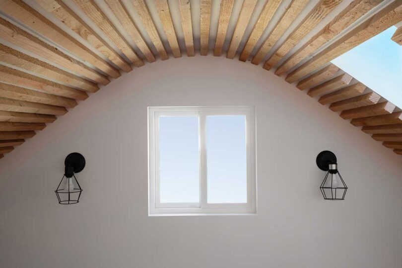interior view of end wall with small window and 2 sconces under pitched roof clad in wood beams