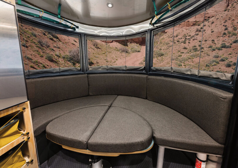 Interior of the the REI Co-op Special Edition Basecamp 20X travel trailer dining area with table covered with cushions to convert into a lounge with panoramic window view.