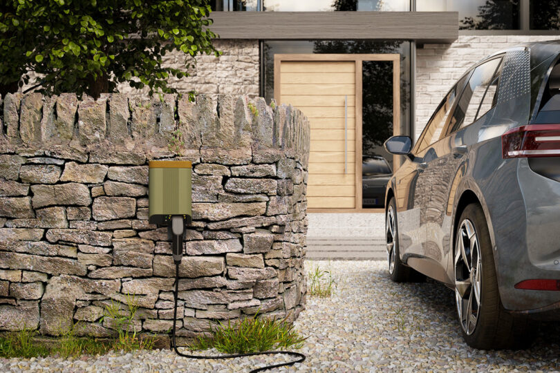 Simpson & Partners EV charging system installed outdoors on a stone wall in front of modern residence and with small hatchback parked and recharging to the left of it.