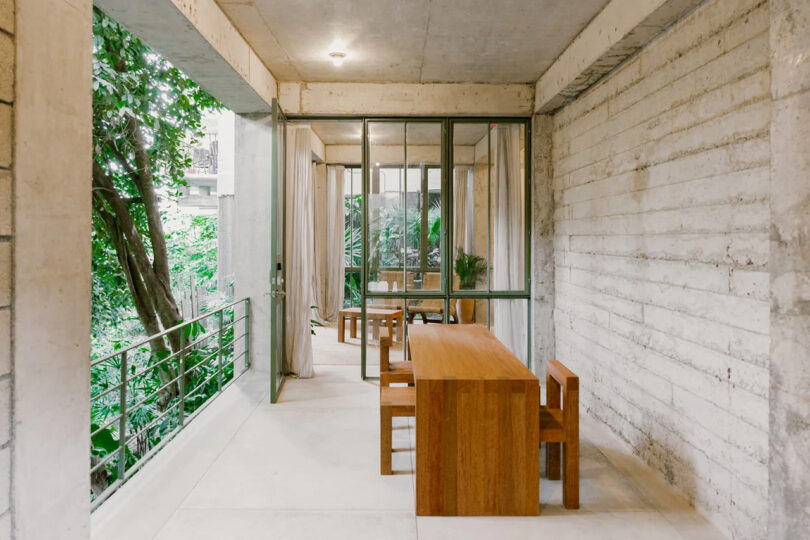 Long narrow wooden dining-console table with two matching dining chairs situated in an indoor-outdoor area with long concrete wall to the right and interior living space visible behind it.