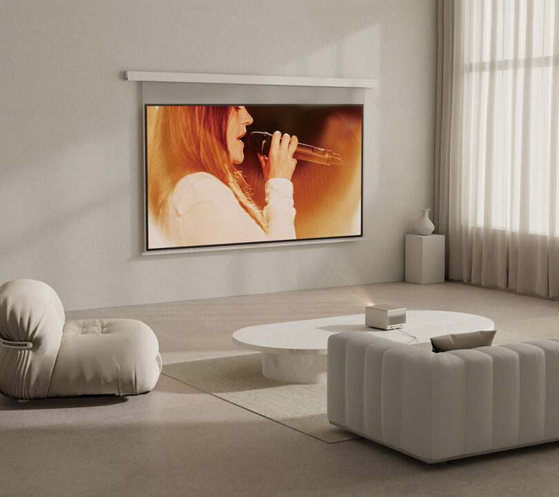 Contemporary living room with low side chair and sofa in off-white and XGIMI HORIZON Ultra protector placed on a low pedestal coffee table. A large projection screen shows a red haired woman from side profile singing into a microphone. Sheer ceiling to floor curtains on the right.