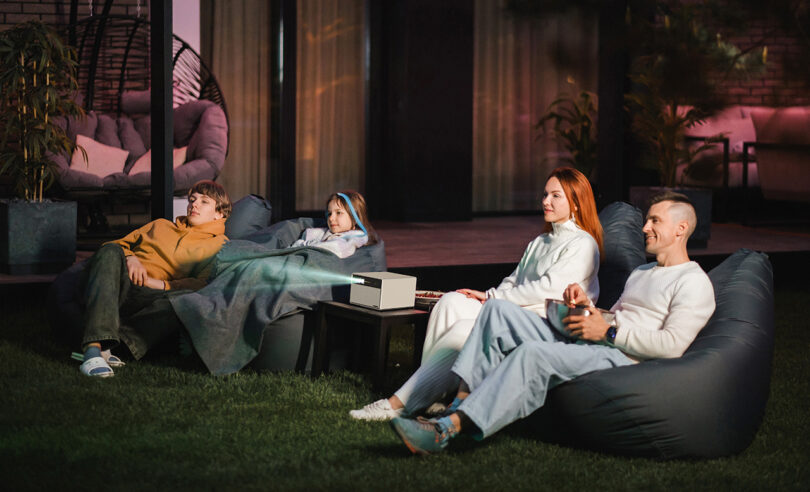 A family of four viewed from the angled front – two adults and young girl and boy – all seated on outdoor beanbags watching a movie in their backyard lawn with the XGIMI HORIZON Ultra projector in the middle.