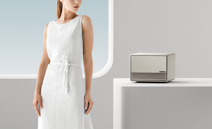 Young white woman in white dress standing the left of the XGIMI HORIZON Ultra hybrid light home projector set upon a white pedestal. A window is in the background with a simulated gradient blue sky.