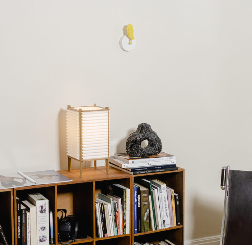 Birdie yellow bird-shaped air quality monitor installed onto wall over low 2 shelf bookcase with small rectangular paper lantern with books and magazines stored in shelves.