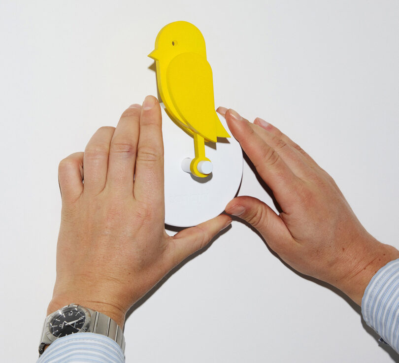 Two hands holding Birdie yellow bird-shaped air quality monitor while installing it onto a white wall.